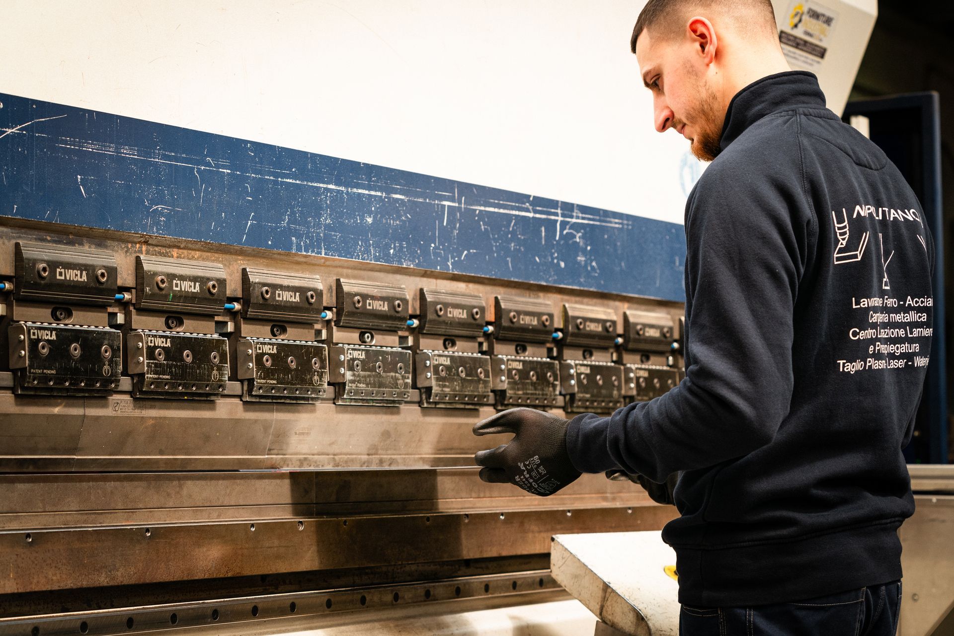 Uomo che aziona una macchina piegatrice per metalli in una fabbrica, indossando guanti e una felpa scura.