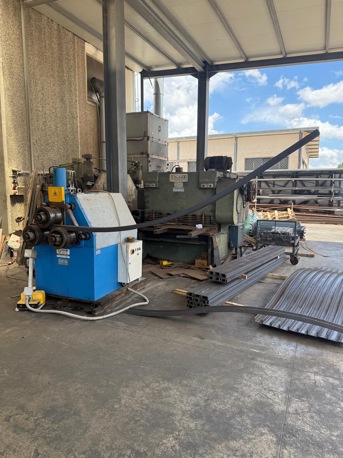 Un macchinario per la lavorazione dei metalli blu e un macchinario industriale verde scuro in un'officina. Sul pavimento ci sono barre di metallo e una lamiera ondulata.