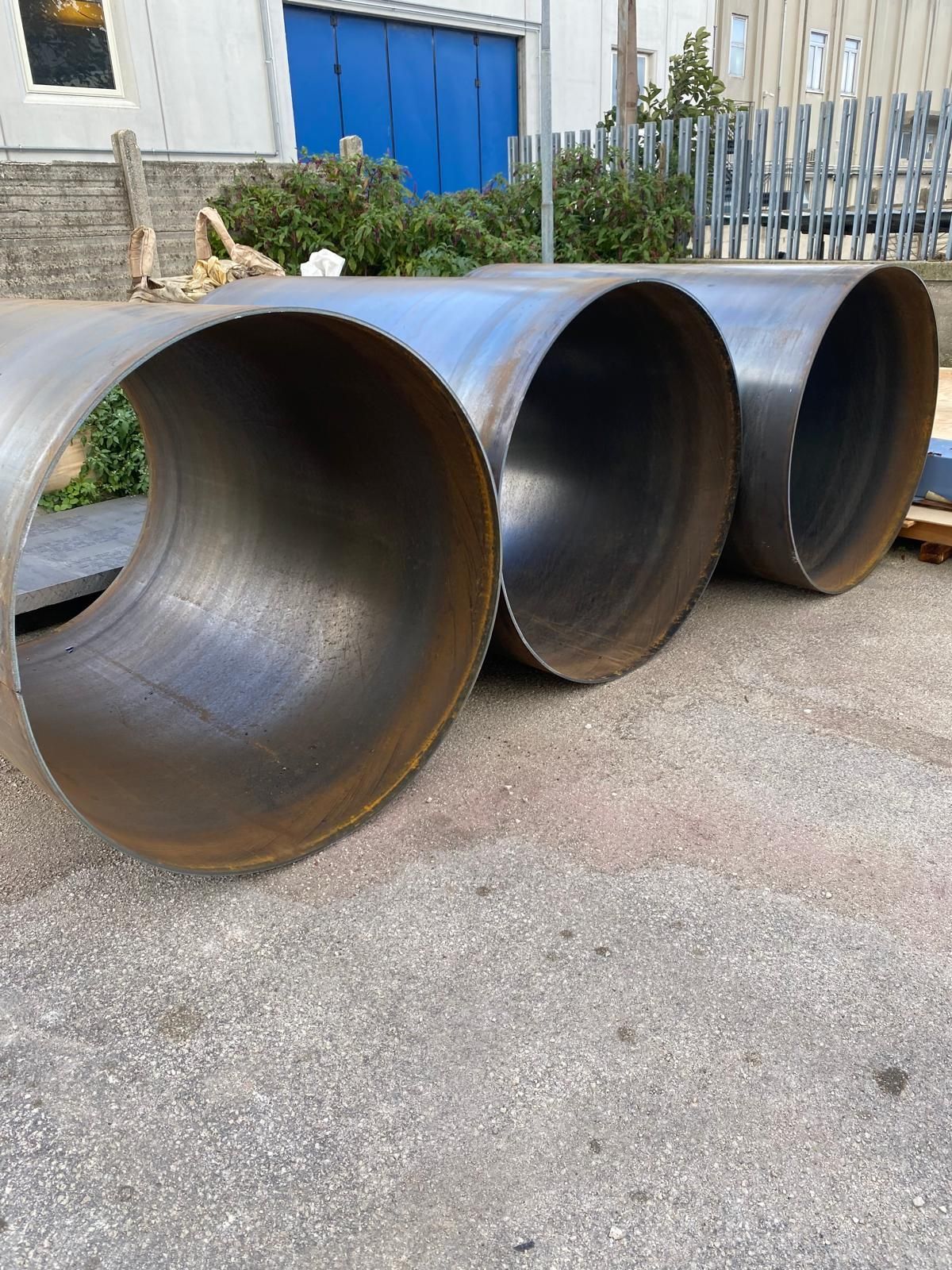 Tre grandi tubi metallici cilindrici appoggiati sul cemento all'esterno di un edificio con porte blu.