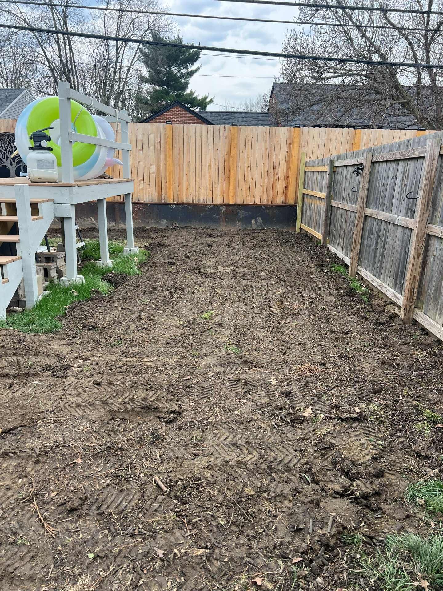 Narrow backyard with bare dirt, wooden fences, and a green fan near a white deck on the left