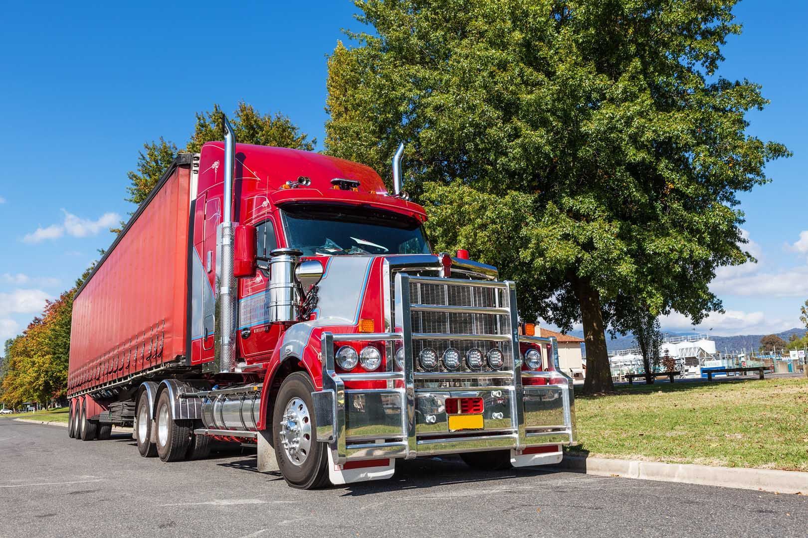 Red semi-truck parked on asphalt road, trees and blue sky in background.