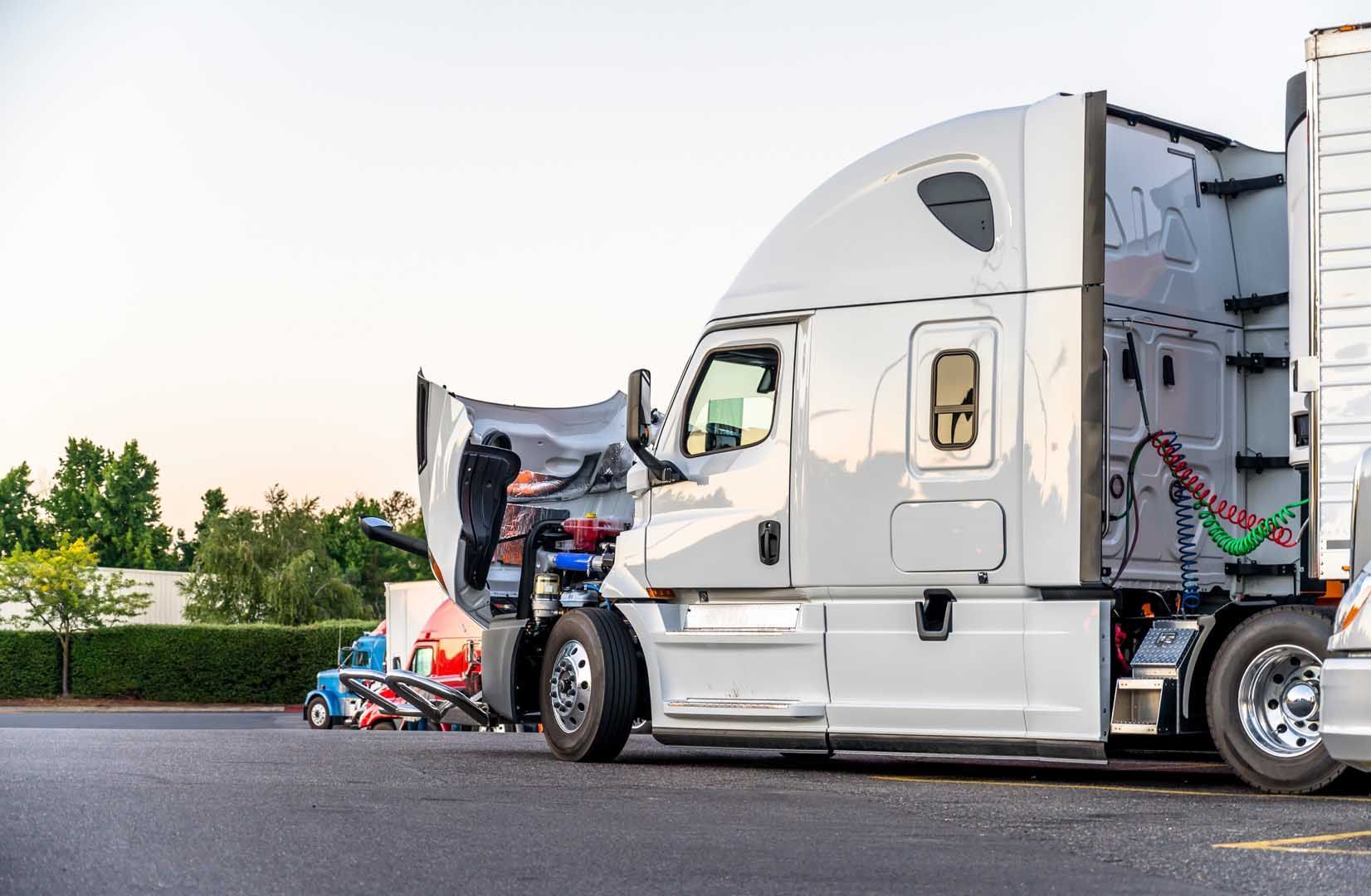 White semi-truck with open hood; parked in a lot. Another truck in the background.