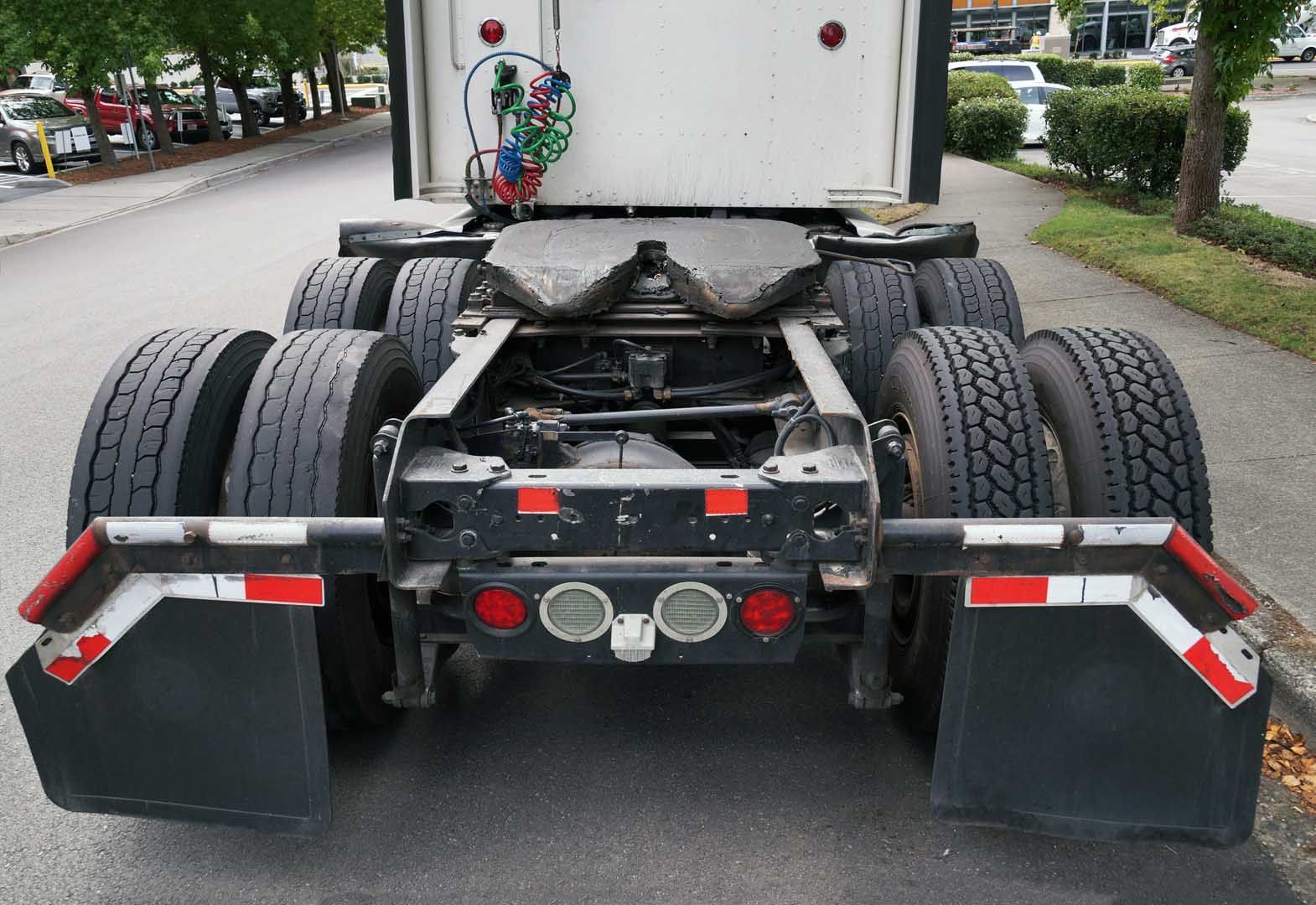 Rear view of a semi-truck chassis and wheels parked on a street; mud flaps with reflective tape.