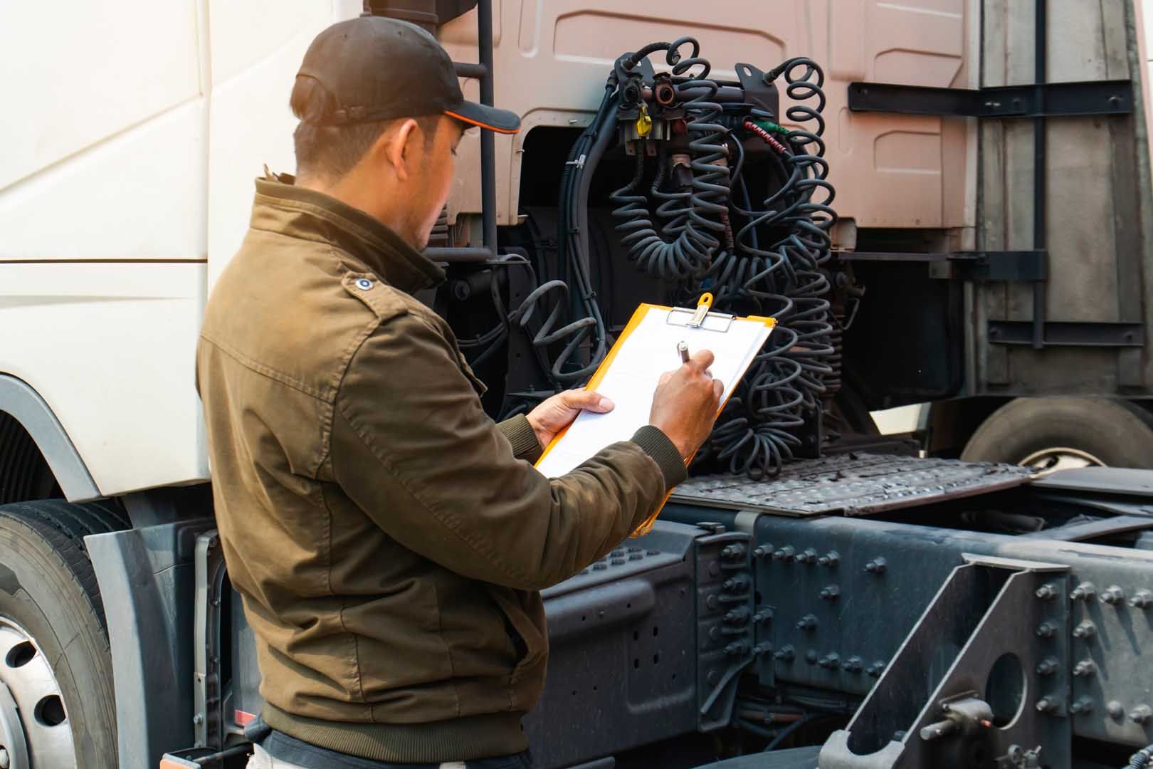 Person in a brown jacket and cap inspecting a truck and writing on a clipboard.