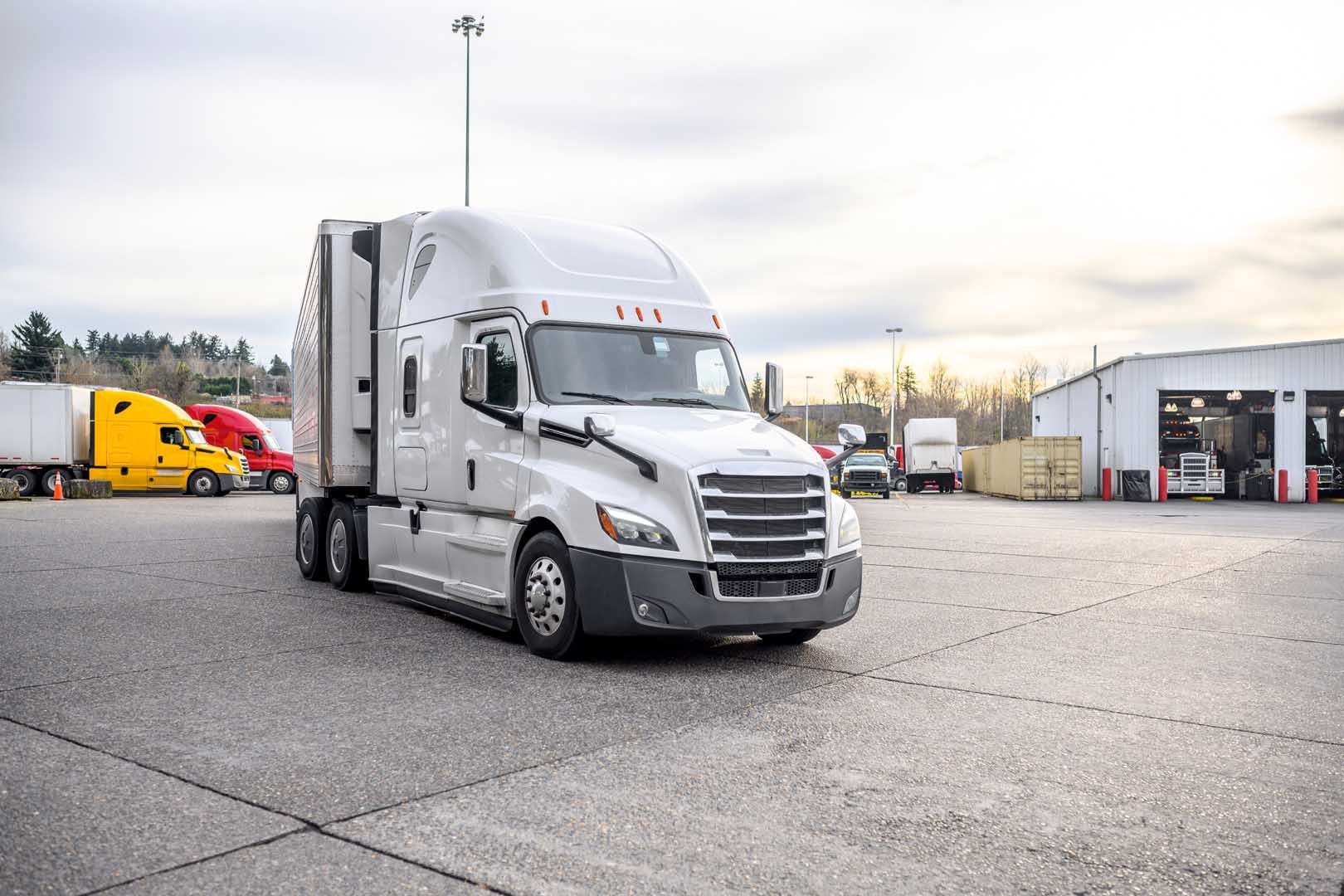 White semi-truck parked in a large lot. Other trucks and a building are in the background under a cloudy sky.