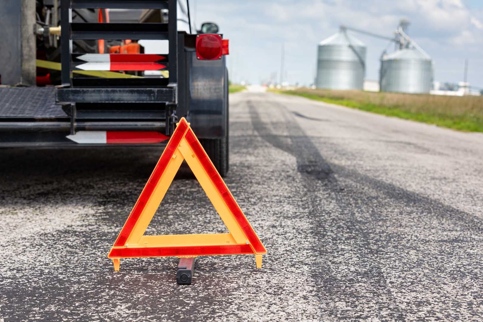 Emergency triangle on road behind a trailer; rural scene with grain silos in the background.