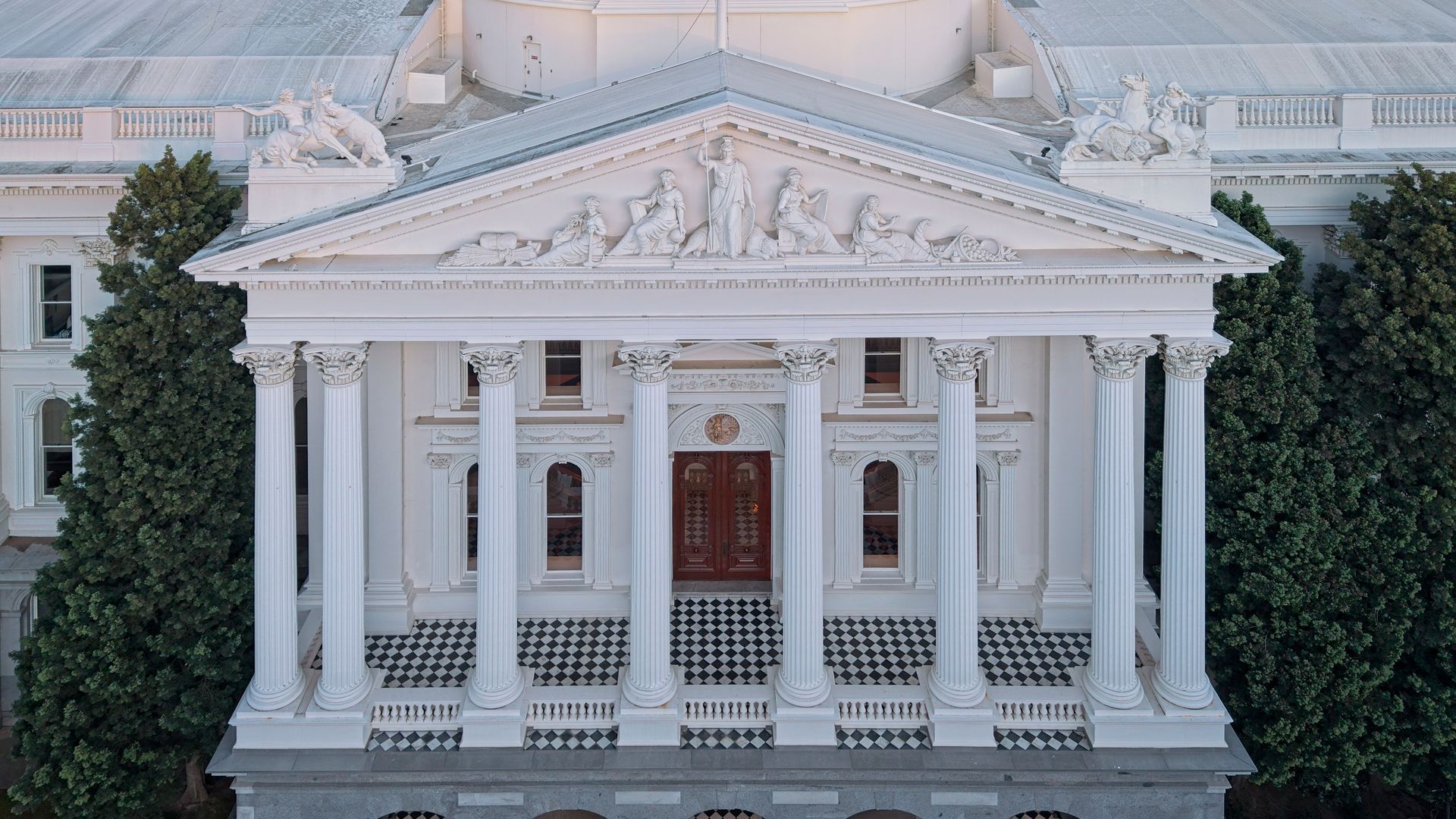 White neoclassical building facade with columns, a triangular pediment, and a central wooden door behind trees