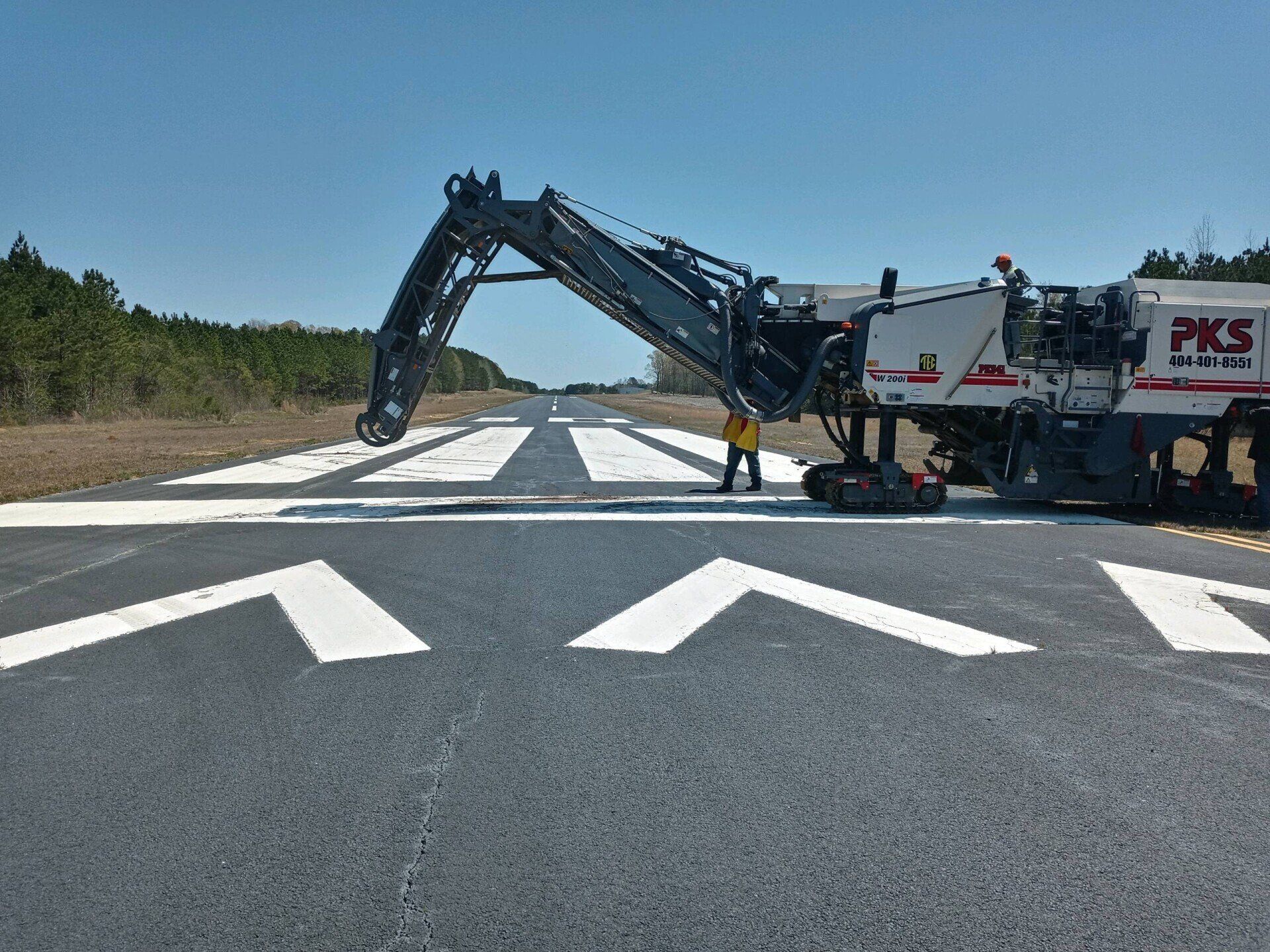 Milling machine on an airport runway; a worker stands nearby, on a sunny day.