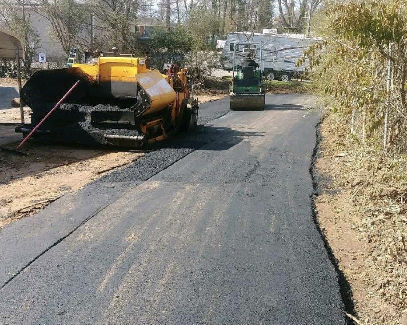 Asphalt paving in progress on a road, with machinery and a roller; a campsite is in the background.