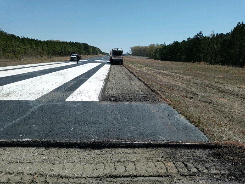Runway with black asphalt patch, white markings, truck, and two people, with trees on both sides.