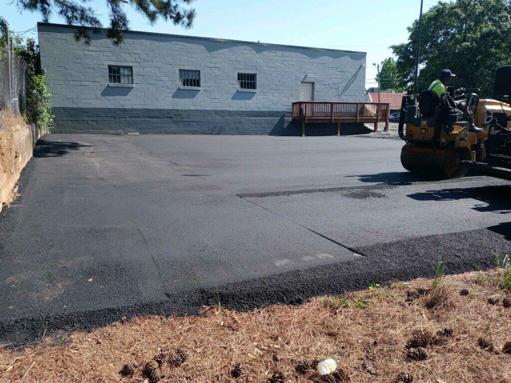Asphalt paving a parking lot; a compact roller works. A building is in the background.