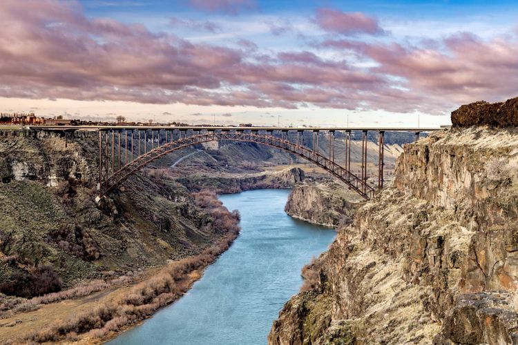 A long, brown bridge spans a deep canyon with a river below. Cloudy sky overhead.