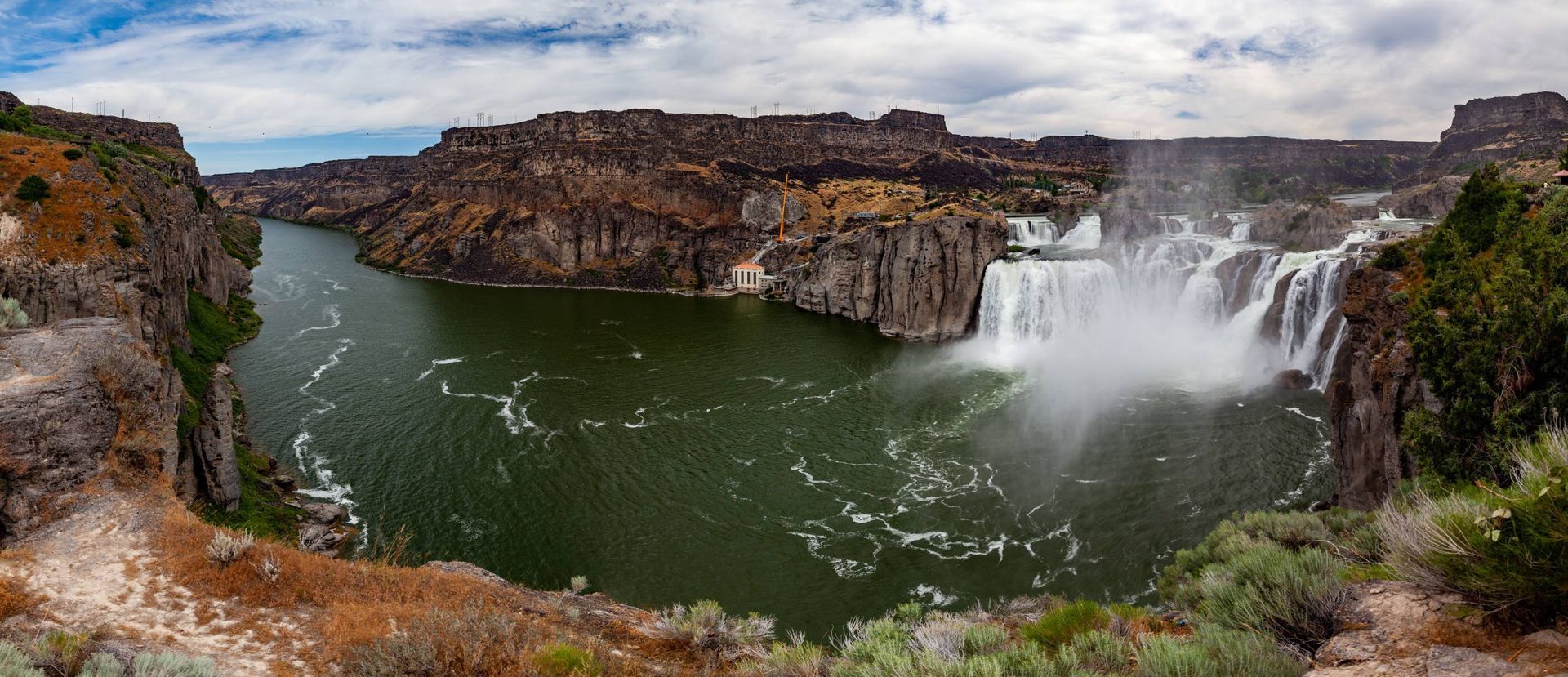 Panoramic view of a waterfall cascading into a green river, surrounded by brown cliffs under a cloudy sky.