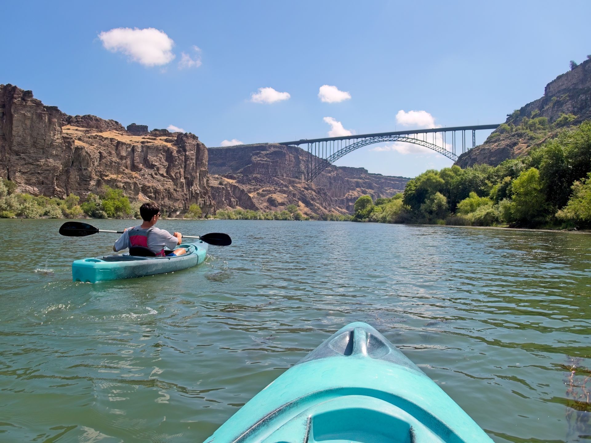 Kayakers on a river under a large bridge, rocky canyon in the background. Sunny day with blue sky.