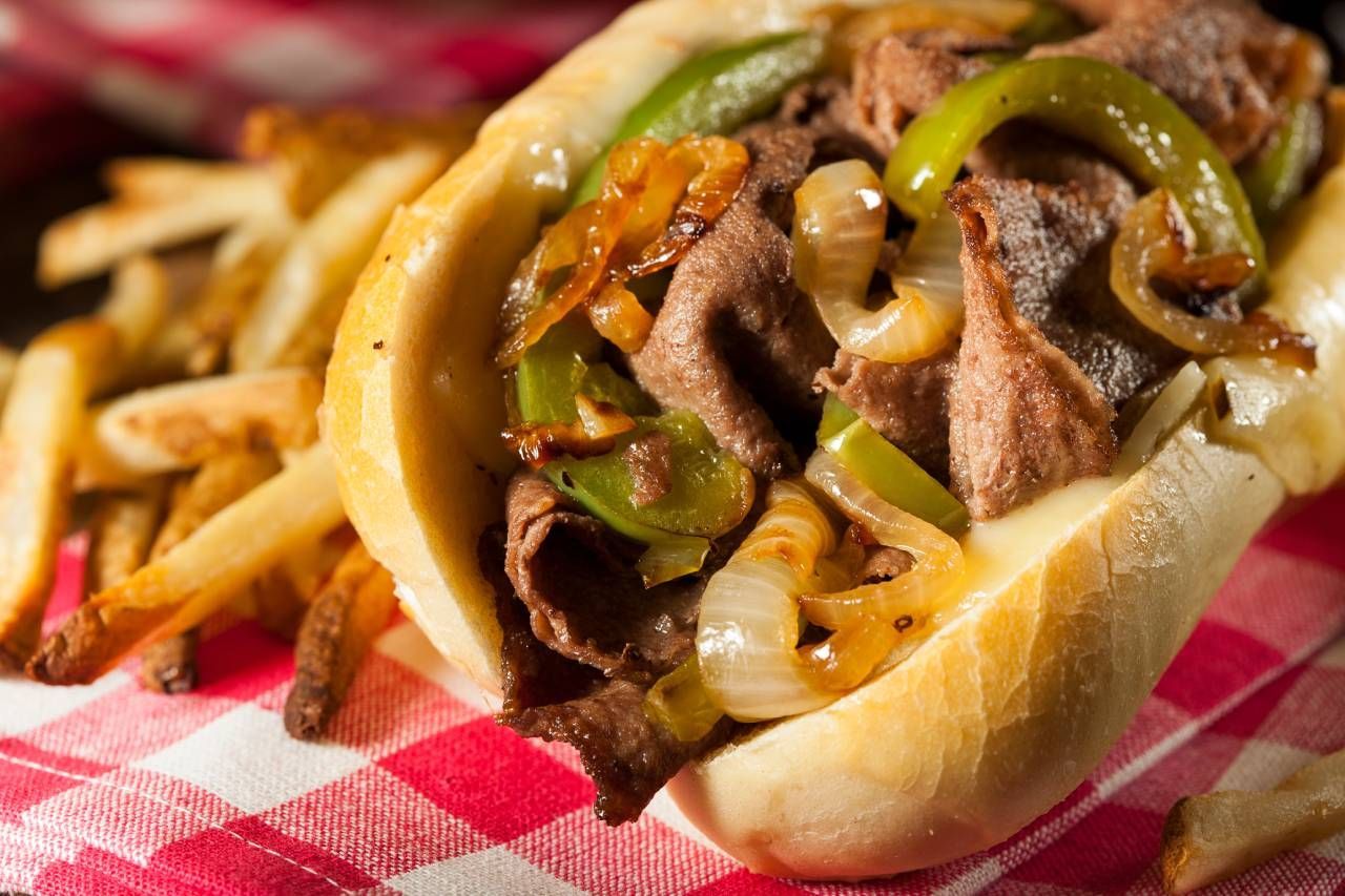 Philly cheesesteak sandwich with peppers and onions next to fries.