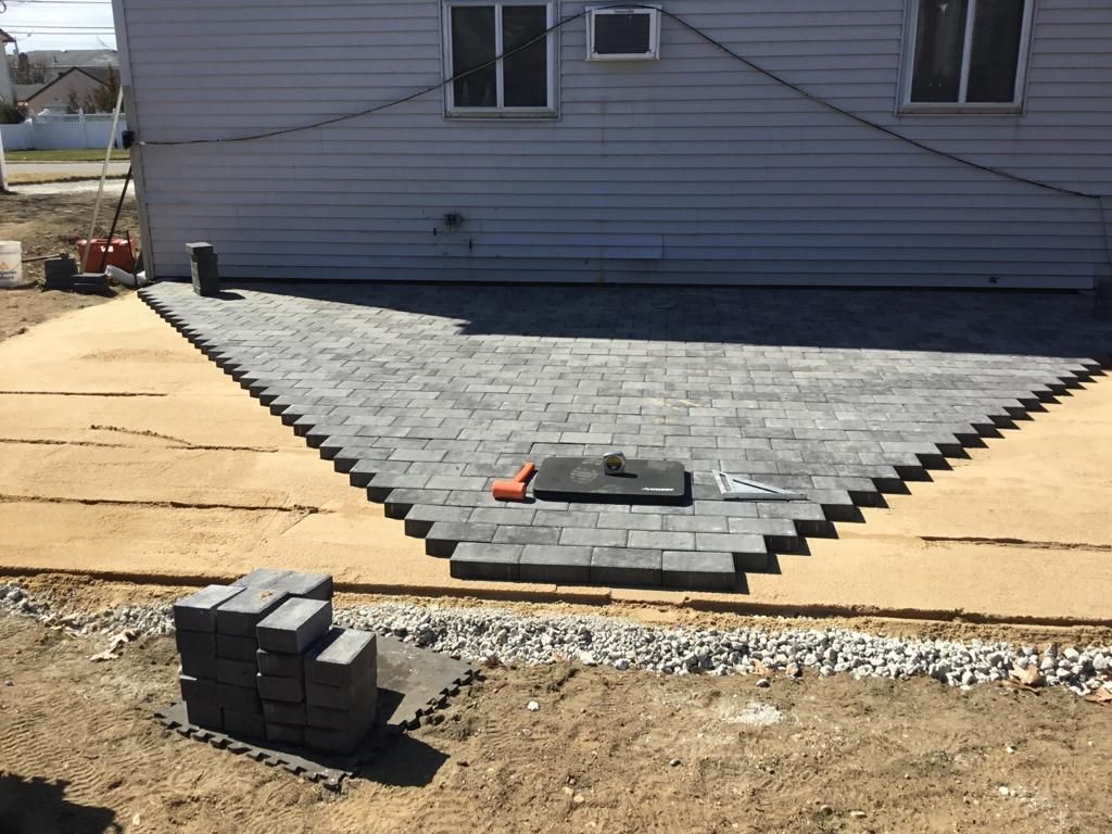 Partially installed gray paver walkway in front of a house, with stacked pavers on a dirt lot.