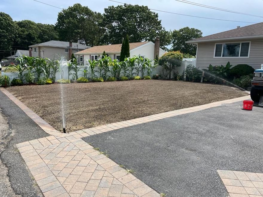 Empty gravel front yard beside a driveway, with a white fence and houses in the background.