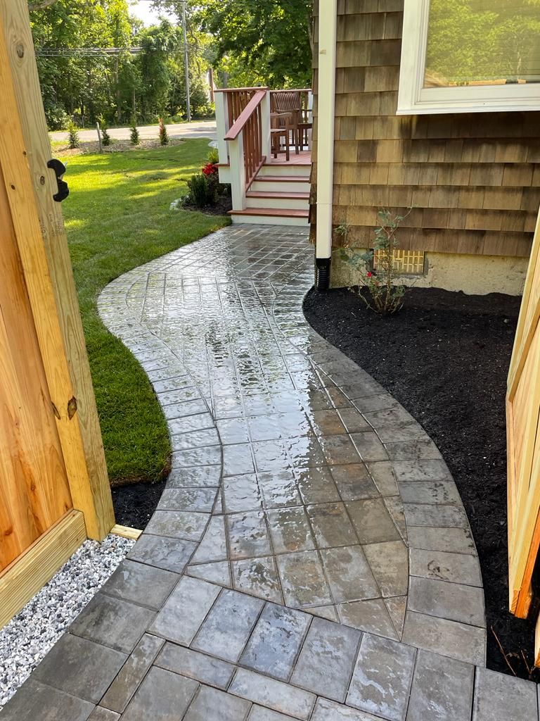 Wet stone walkway beside a house and wooden steps, bordered by gravel and lawn.