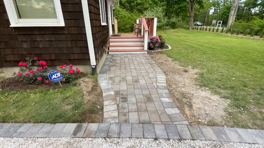 Stone walkway leads to a house porch, bordered by red flowers and a lawn.