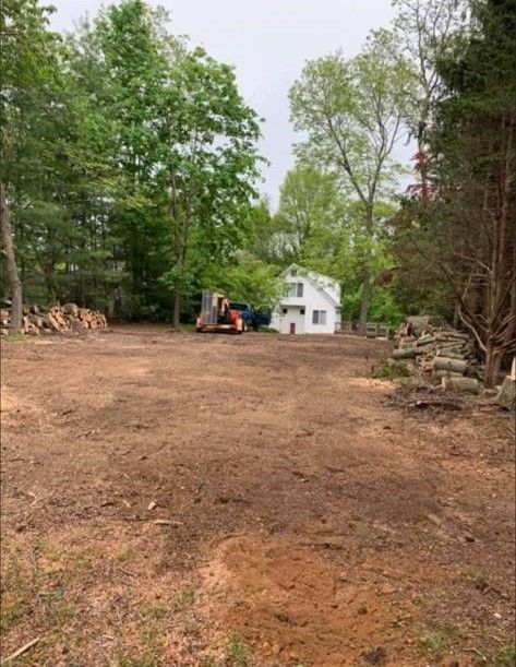 Cleared dirt lot with trees, a small white house in the distance, and an excavator on the left