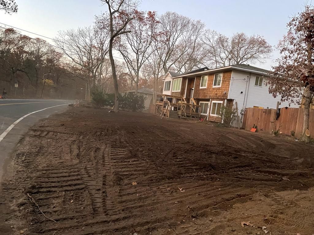 Freshly graded dirt lot beside a curving road and brick house in a wooded neighborhood