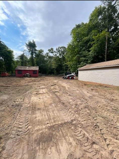 Cleared dirt lot beside a white garage and red shed, with trees under a cloudy sky.