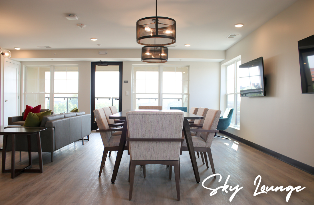 Sky Lounge interior with dining table, seating, and lighting. Gray walls, windows, and light wood flooring.