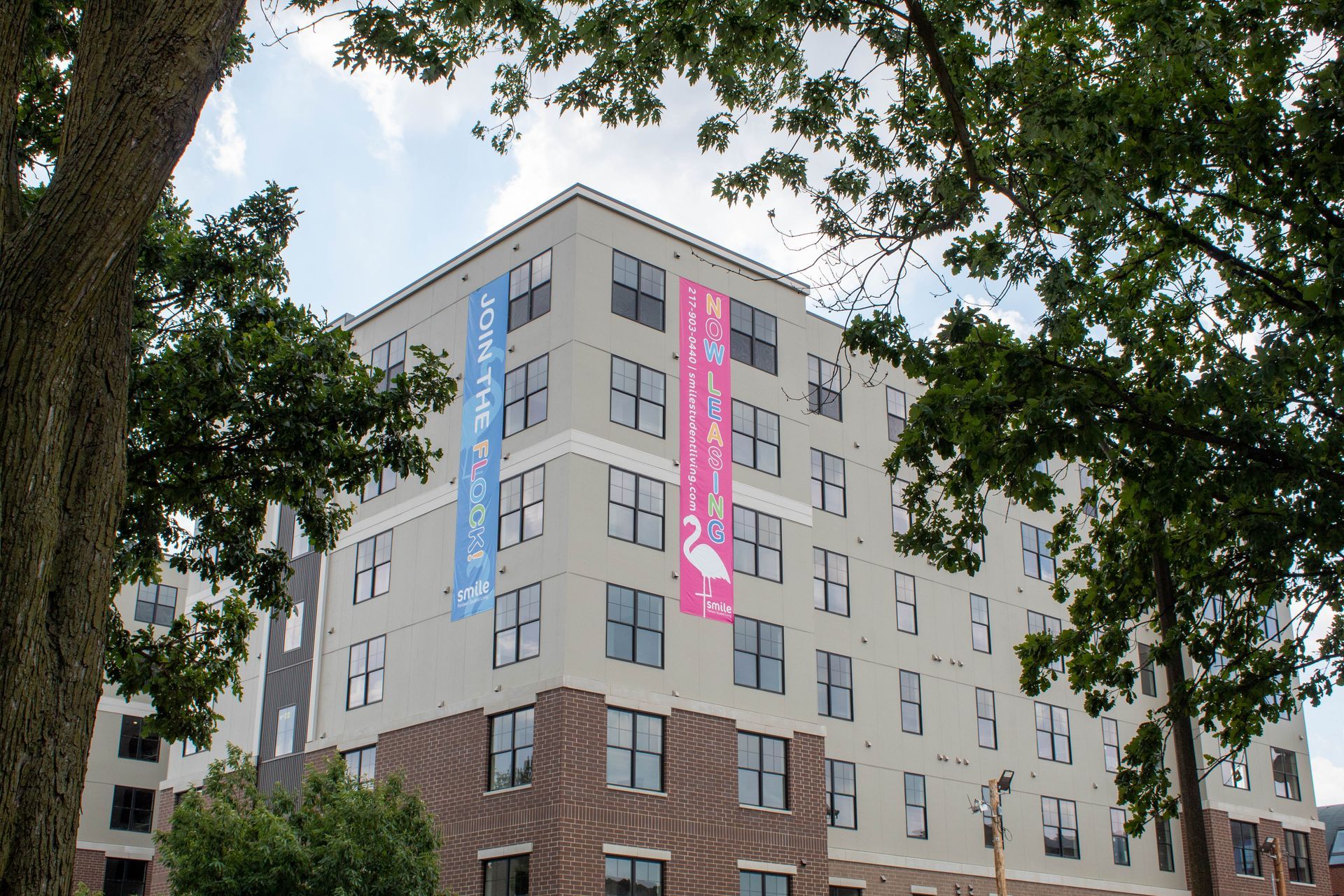 Multi-story apartment building with banners, seen through trees, against a cloudy sky.