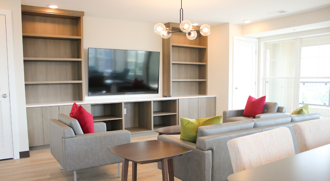 Living room with built-in shelves, TV, gray sofas, colorful pillows, and a small brown table. Bright with natural light.