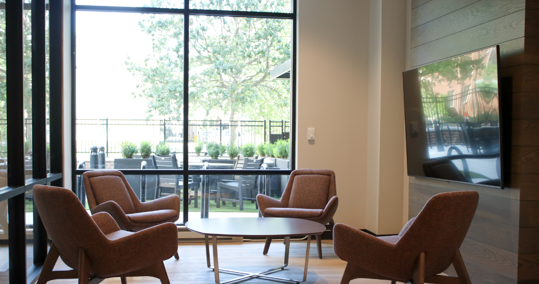 Four brown chairs arranged around a table in a lobby with a window overlooking an outdoor patio.