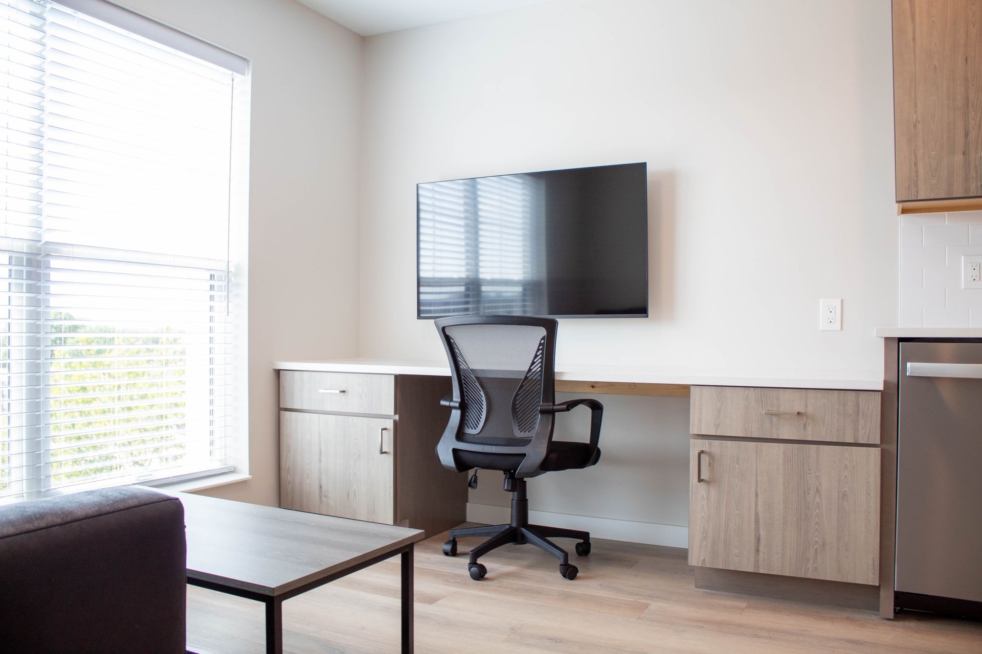 A modern room with a desk, chair, TV, window, and small kitchen area.