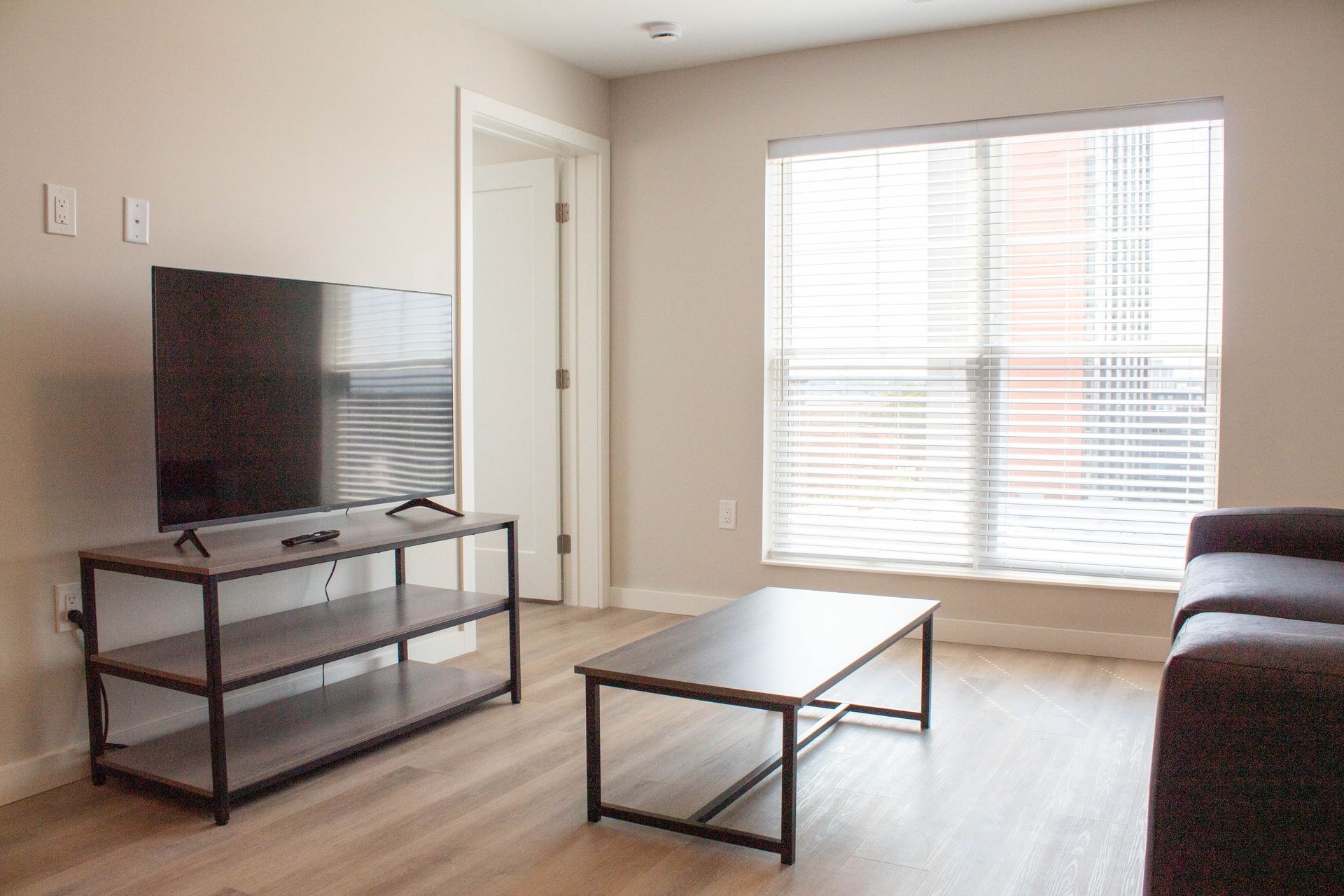 Living room with TV, window with blinds, coffee table, and couch. Beige walls, light wood flooring.
