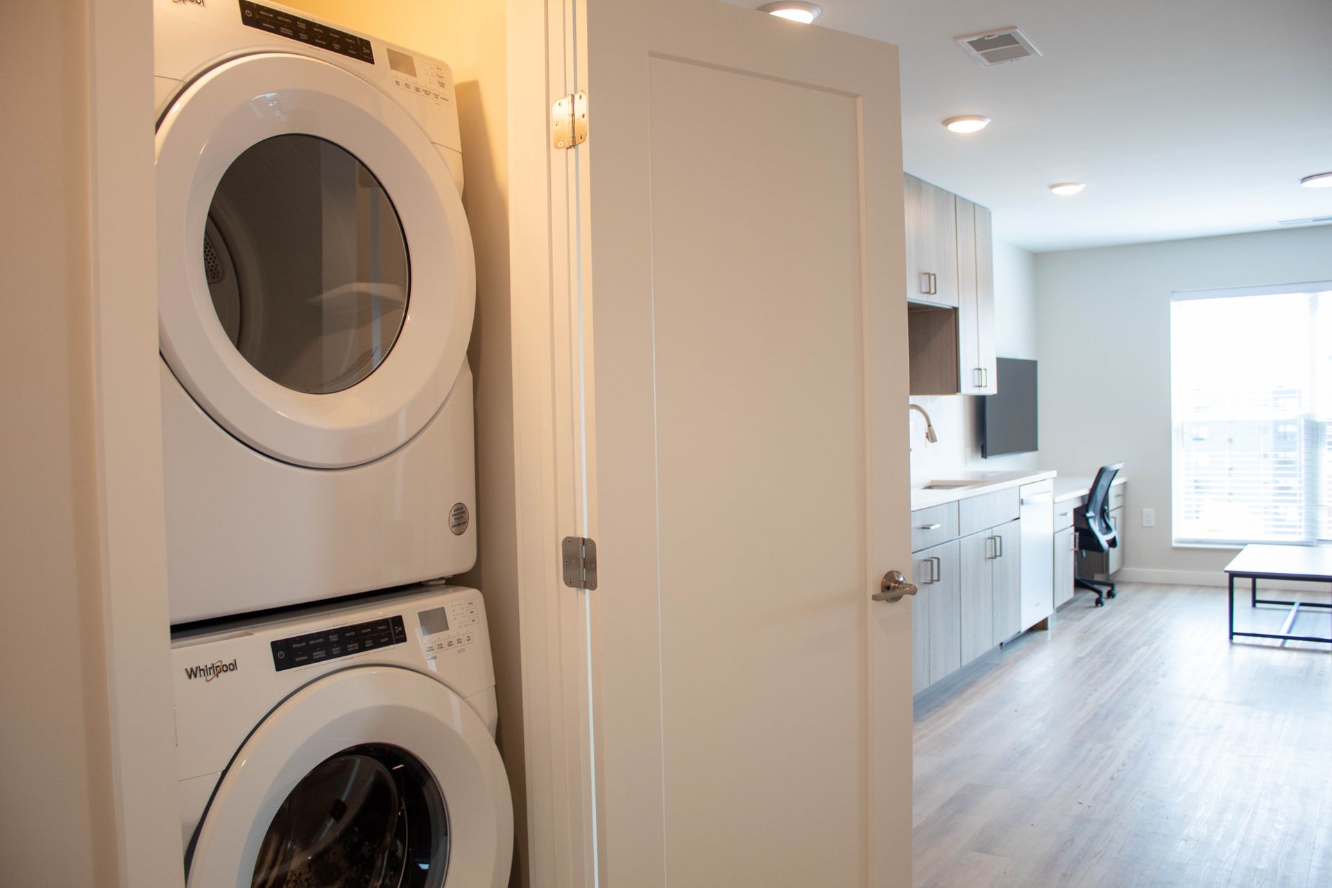 Stacked white washer and dryer in a small laundry area, door open to living space with desk.