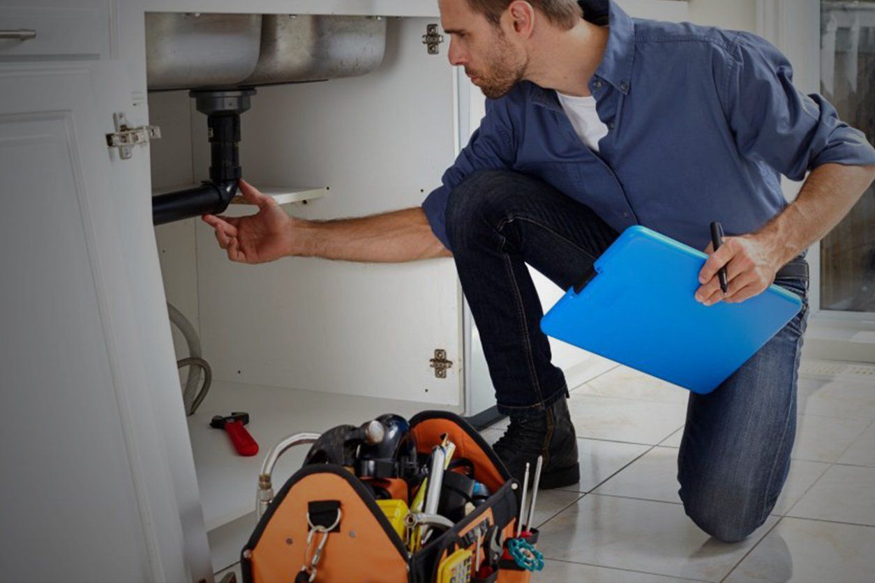 A man is kneeling down under a sink while holding a clipboard.
