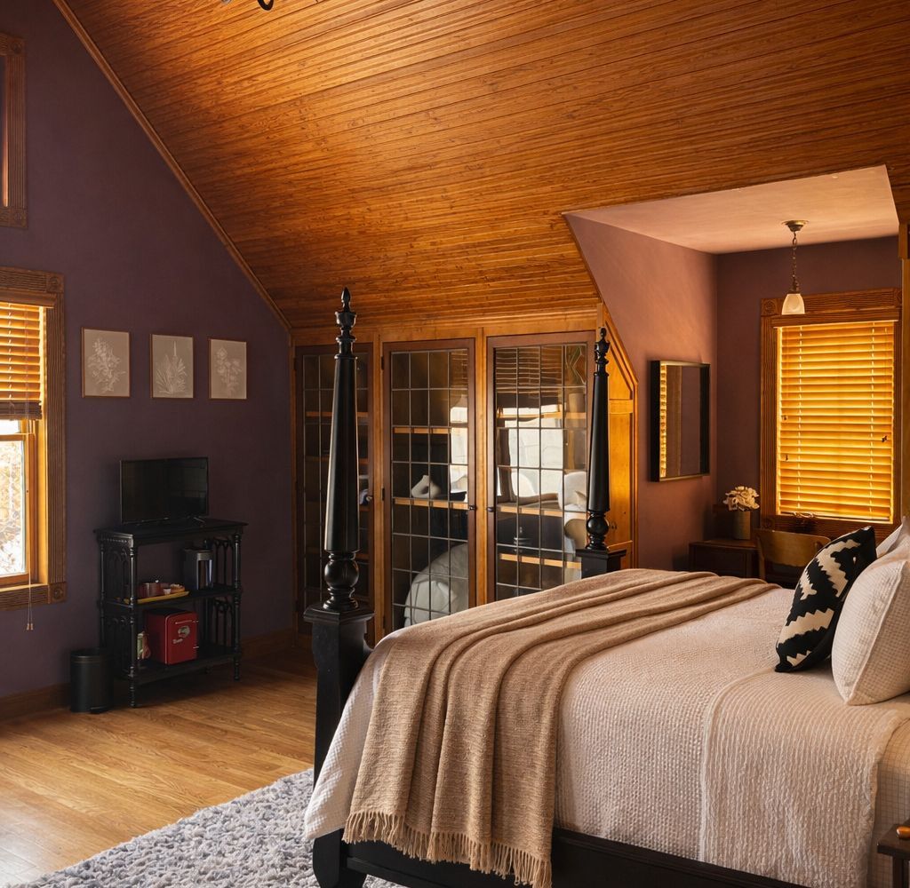 A bedroom with a dark wooden bed, purple walls, a wood-paneled sloped ceiling, an ornate chandelier, and a textured rug.