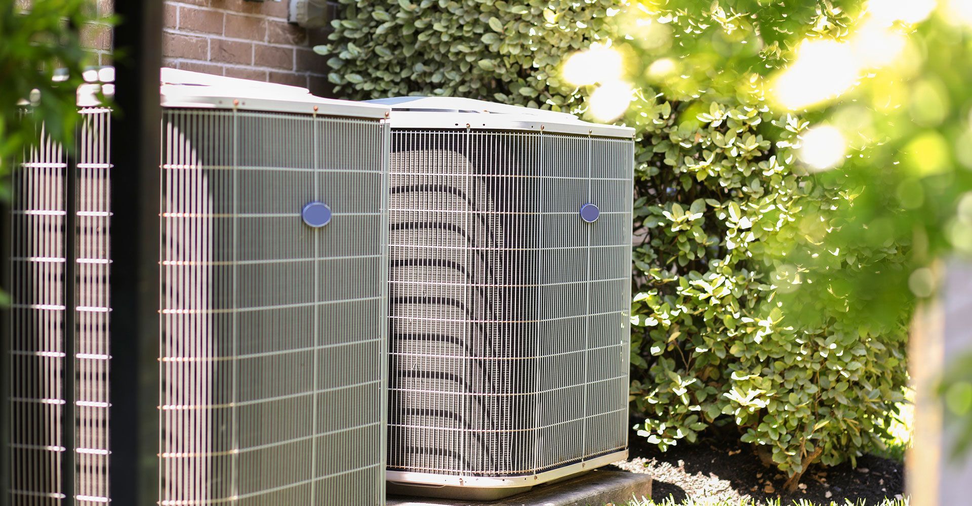 Three air conditioning units next to a brick wall, surrounded by green bushes and trees.
