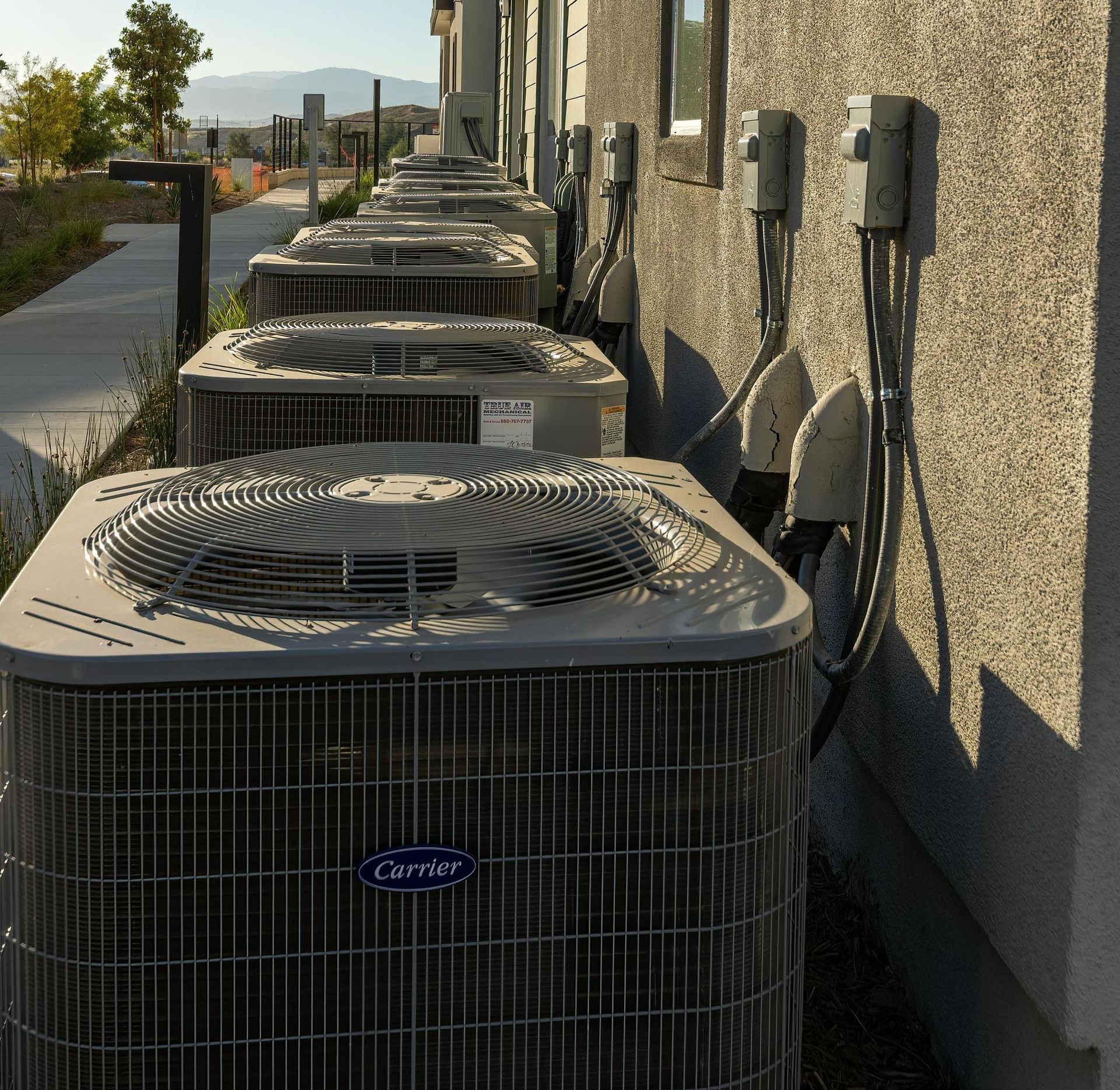 Row of gray Carrier air conditioning units along a building, with electrical boxes on the wall.