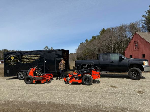 Man stands with lawn equipment and black truck by a black trailer and red barn.