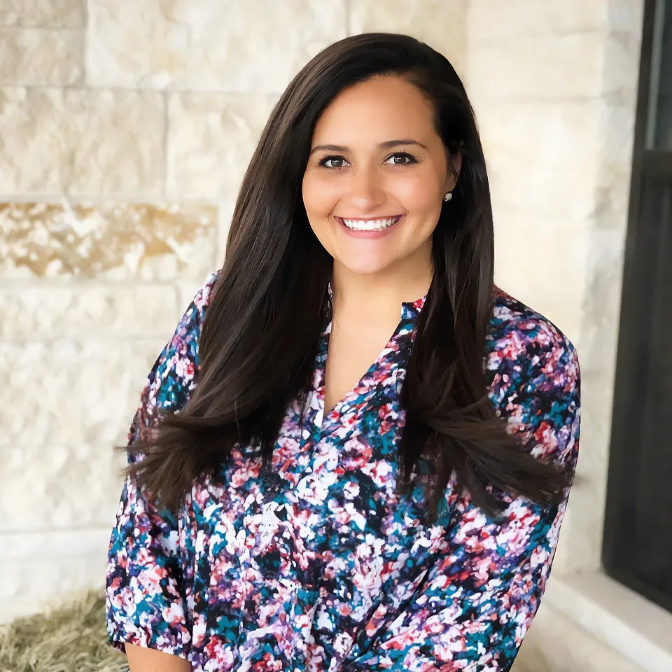 Woman with long dark hair smiles, wearing a floral top, standing near a stone wall.