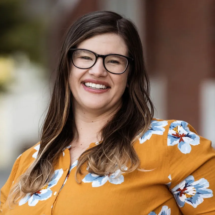 Woman wearing glasses smiles at the camera, in a yellow floral top. Blurred urban background.