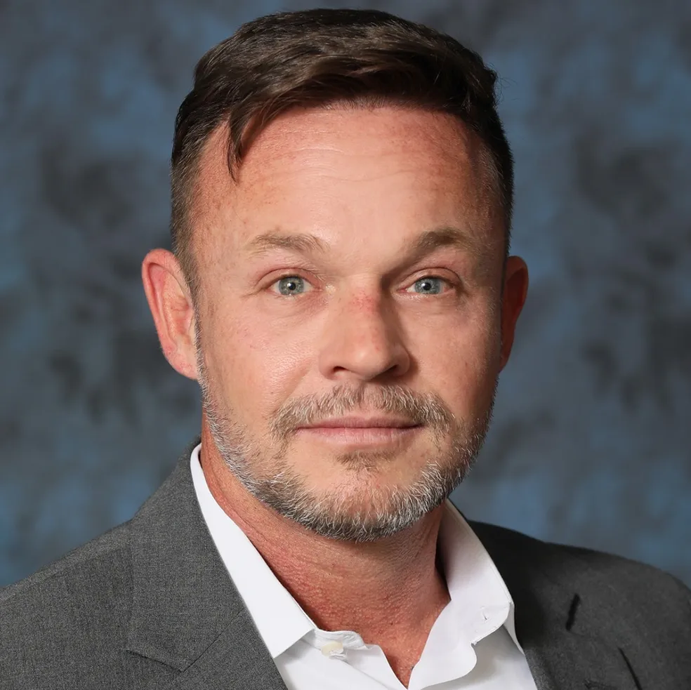 Man in a gray suit jacket and white shirt, looking towards the camera with a neutral expression.