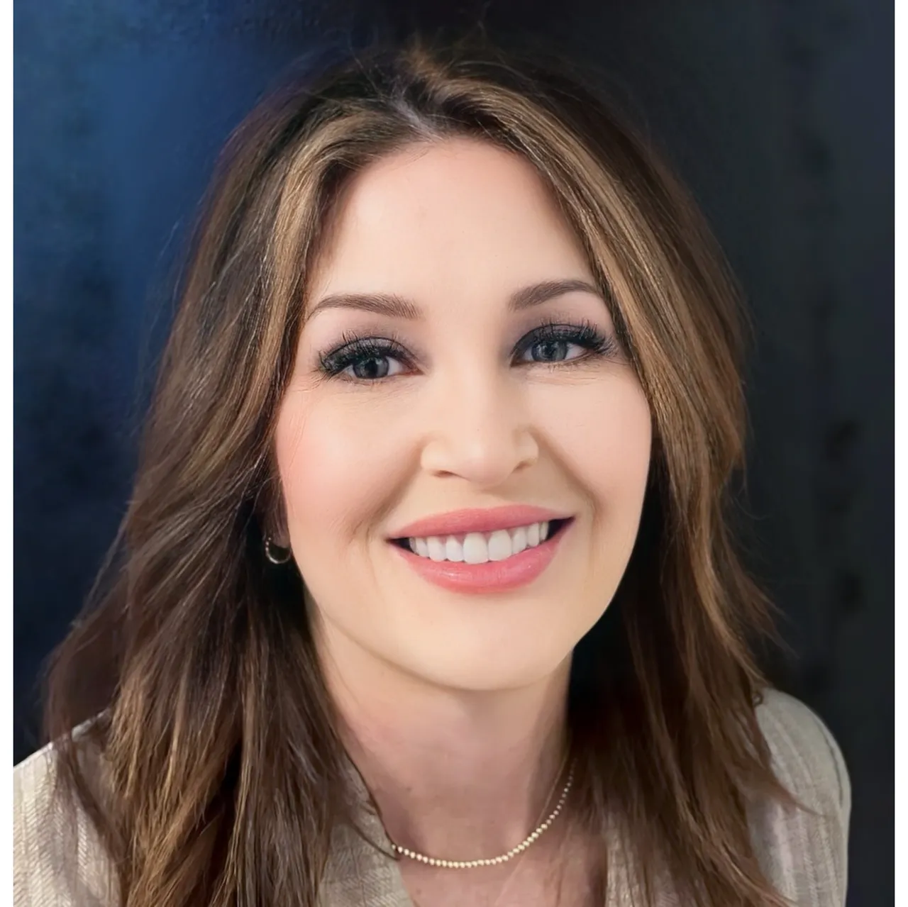 Woman with long brown hair, smiling, wearing a light-colored top, in front of a dark blue backdrop.