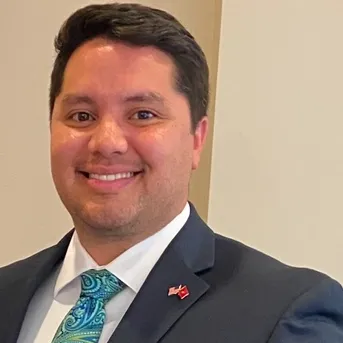 Man in suit smiling, looking at the camera. Teal patterned tie, small red and white pin on lapel.