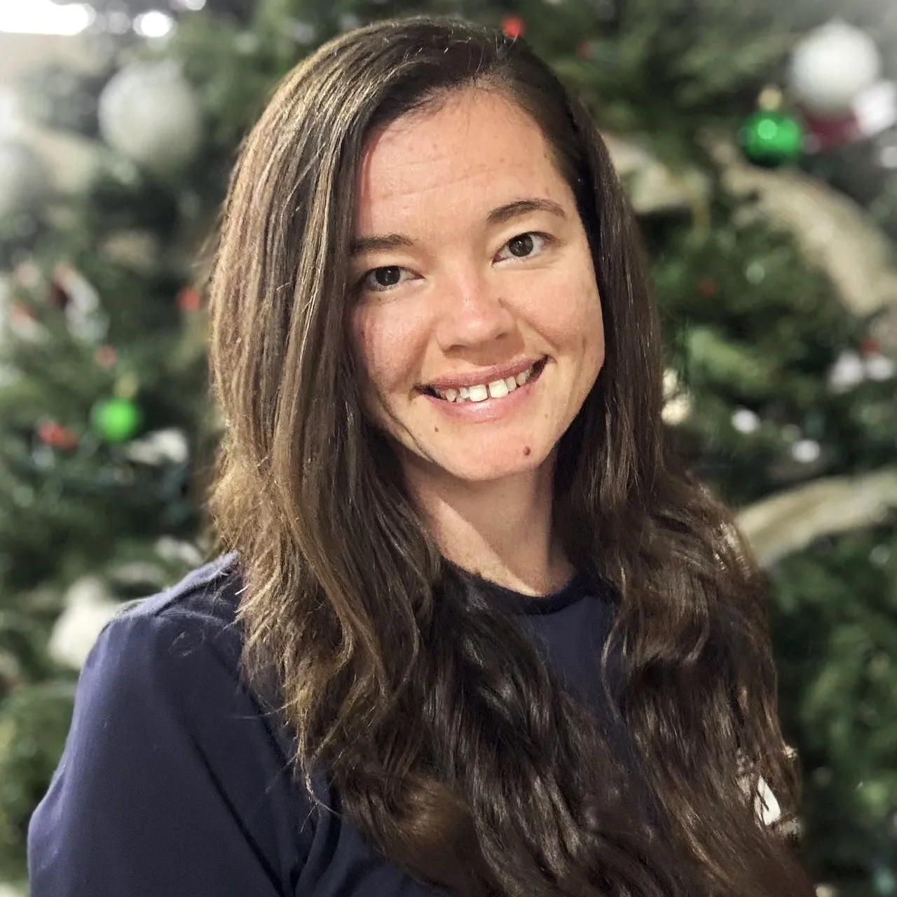 Woman with long brown hair smiles in front of a Christmas tree, wearing a dark blue shirt.