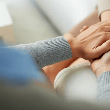 Hands clasped together, one hand on top of another. A person in a gray sweater comforts another.