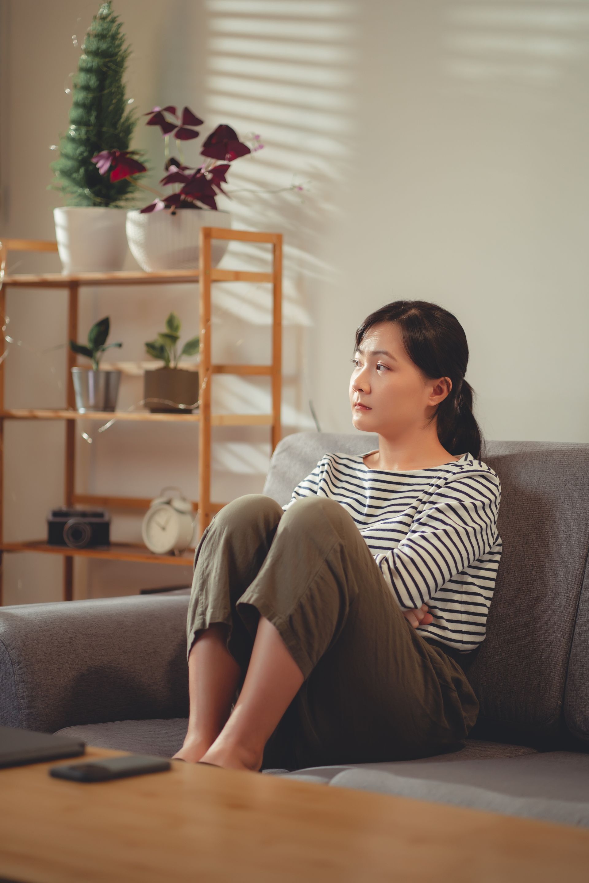 Woman sitting on couch, arms crossed, looking away. Living room setting with sunlight, plants, and phone.