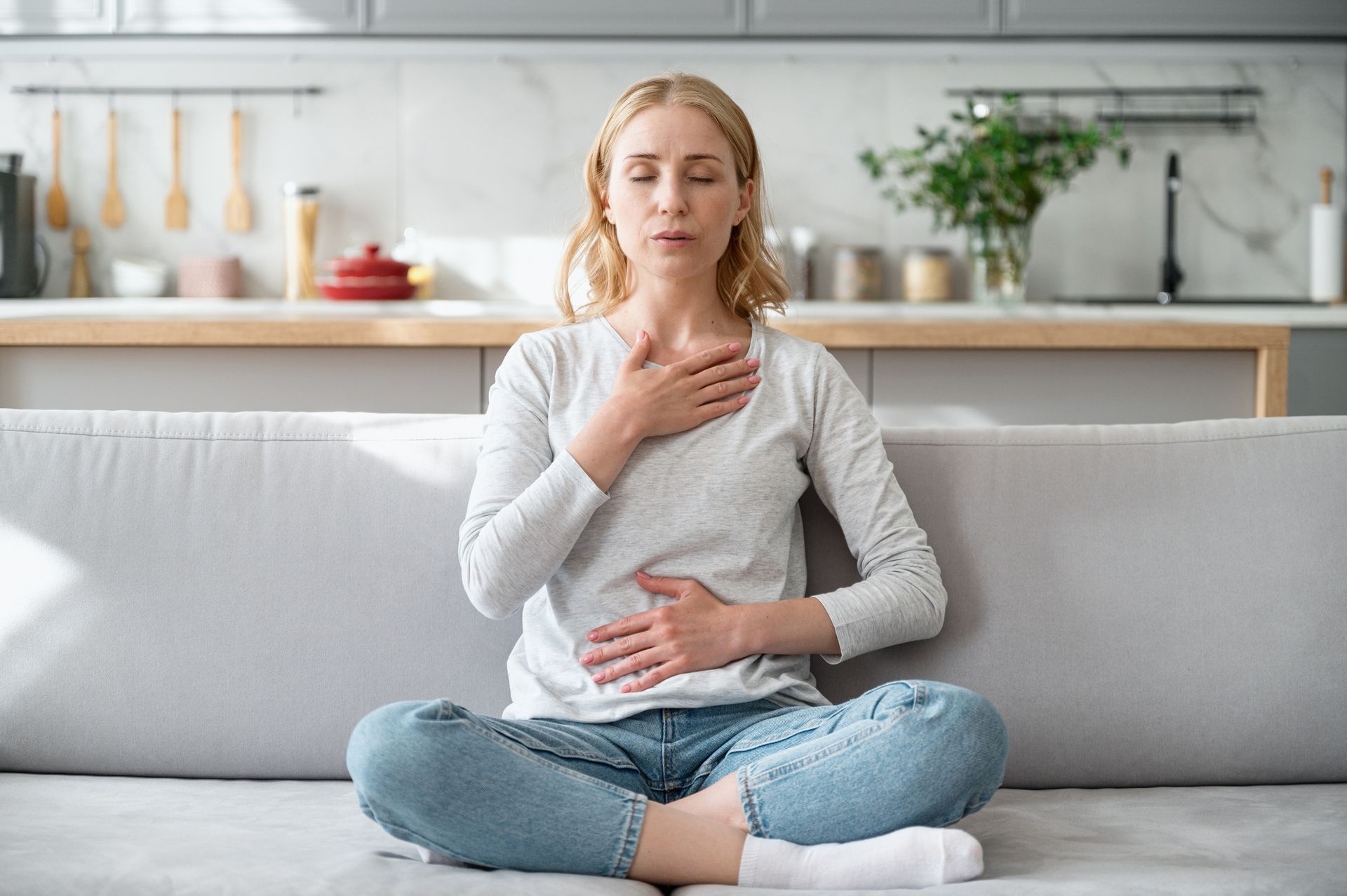 Woman meditating on a sofa, hands on chest and stomach, eyes closed. Interior with plants and kitchen.