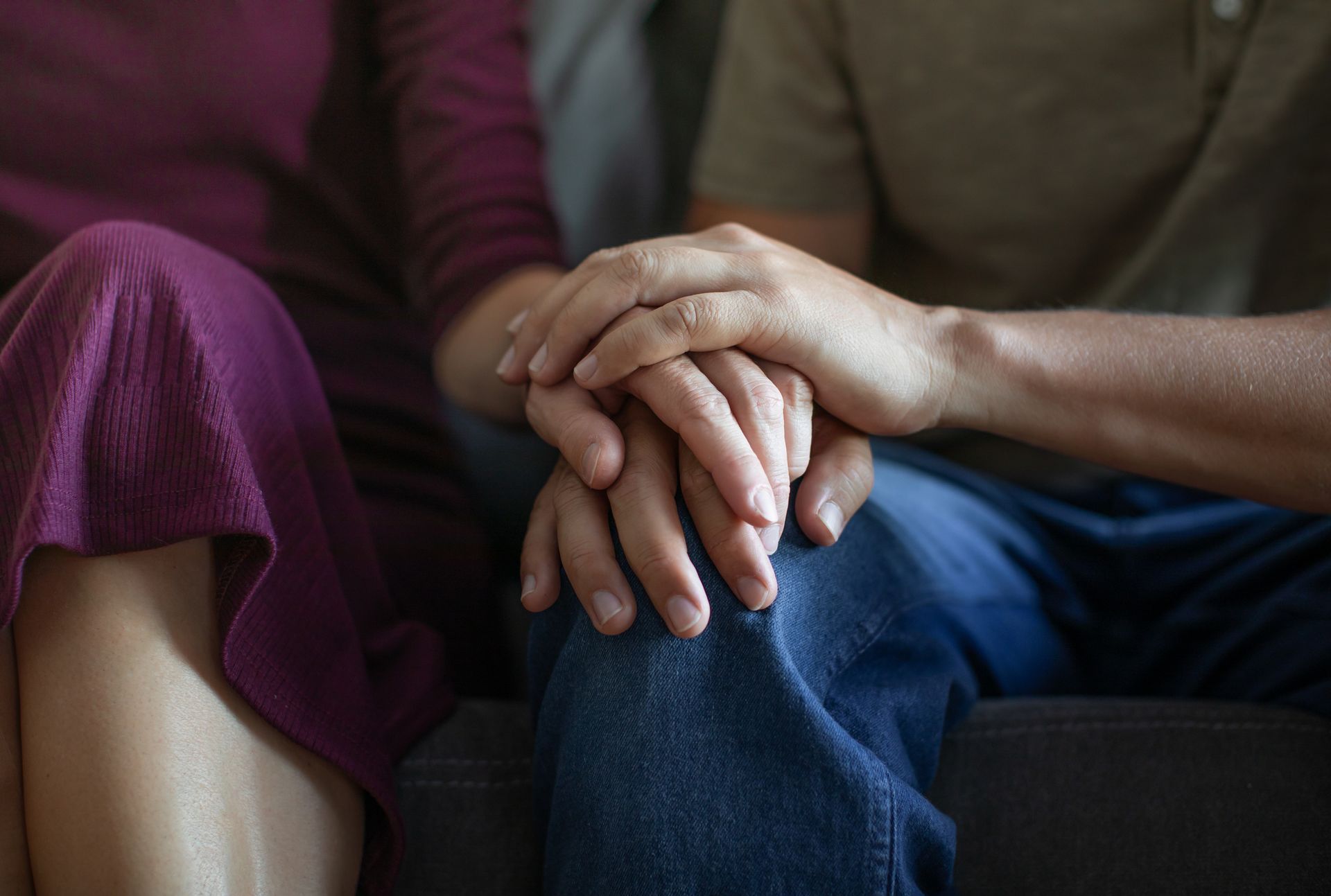 Two people, hands clasped in support, resting on a person's lap; one in purple dress, the other in jeans.