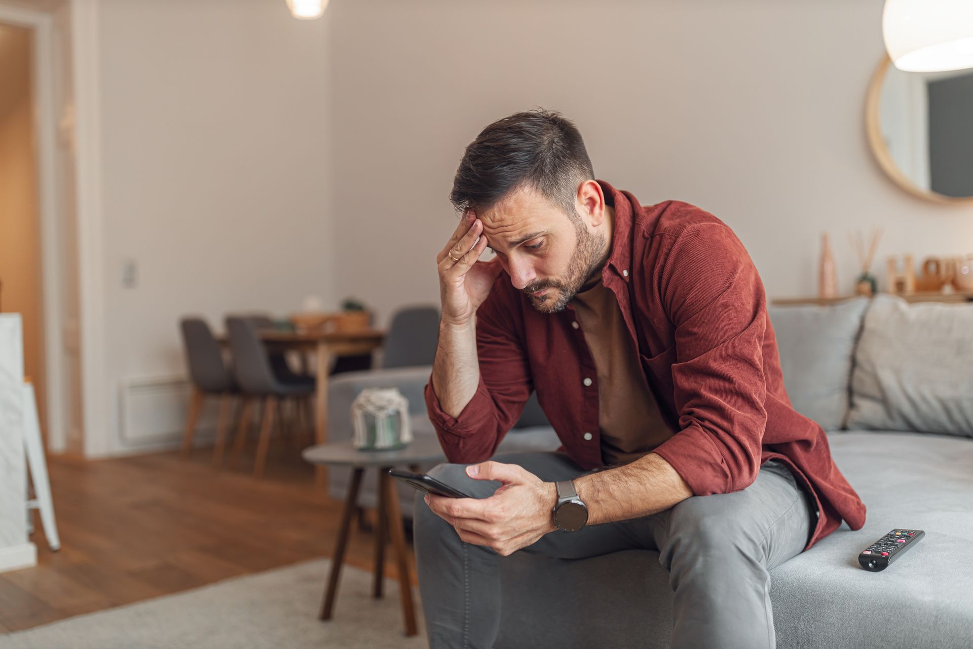 Man sitting on a couch, looking at a phone with a stressed expression; indoors.