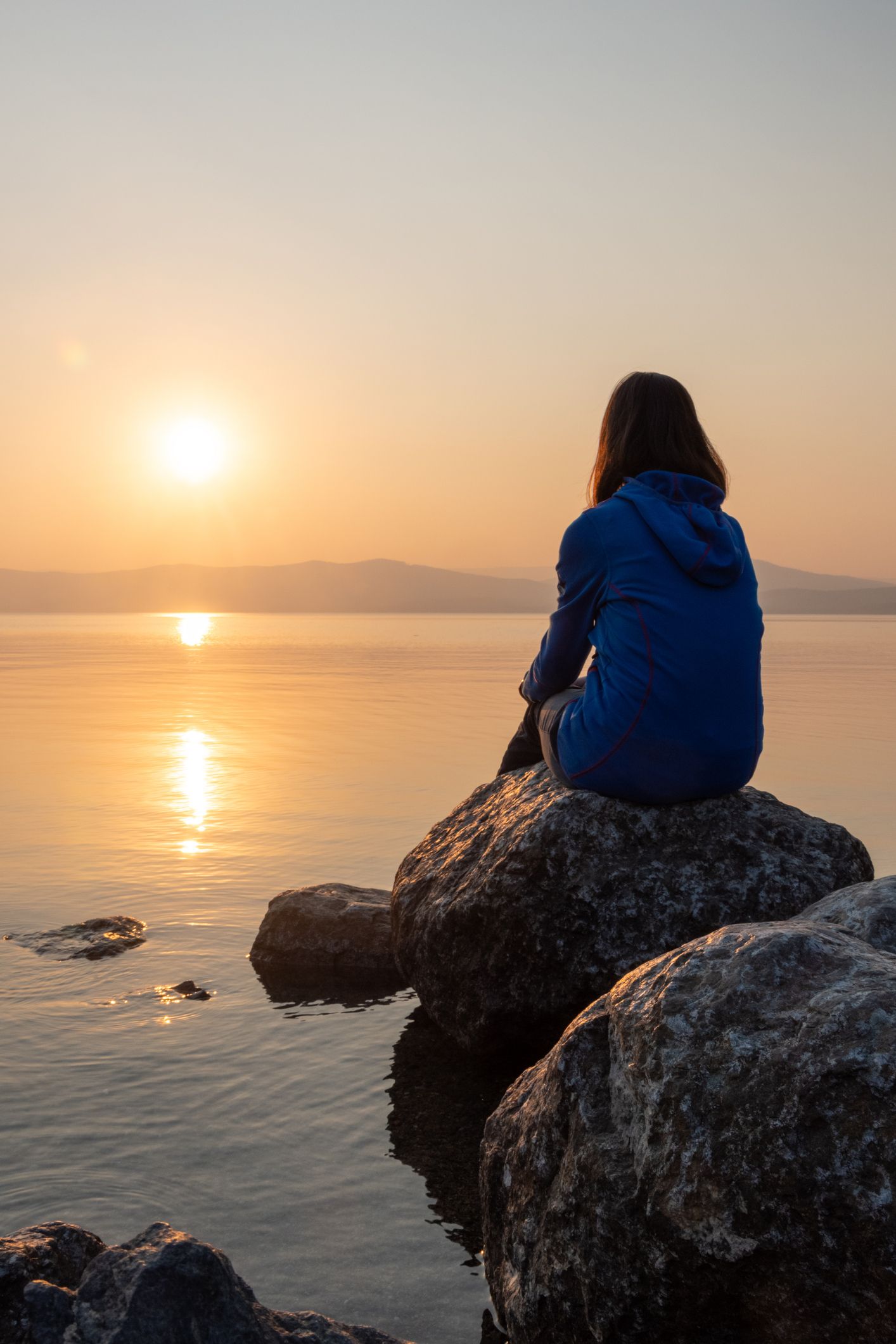 Person sitting on a rock, facing a calm lake at sunset. The sky is golden, person in blue.